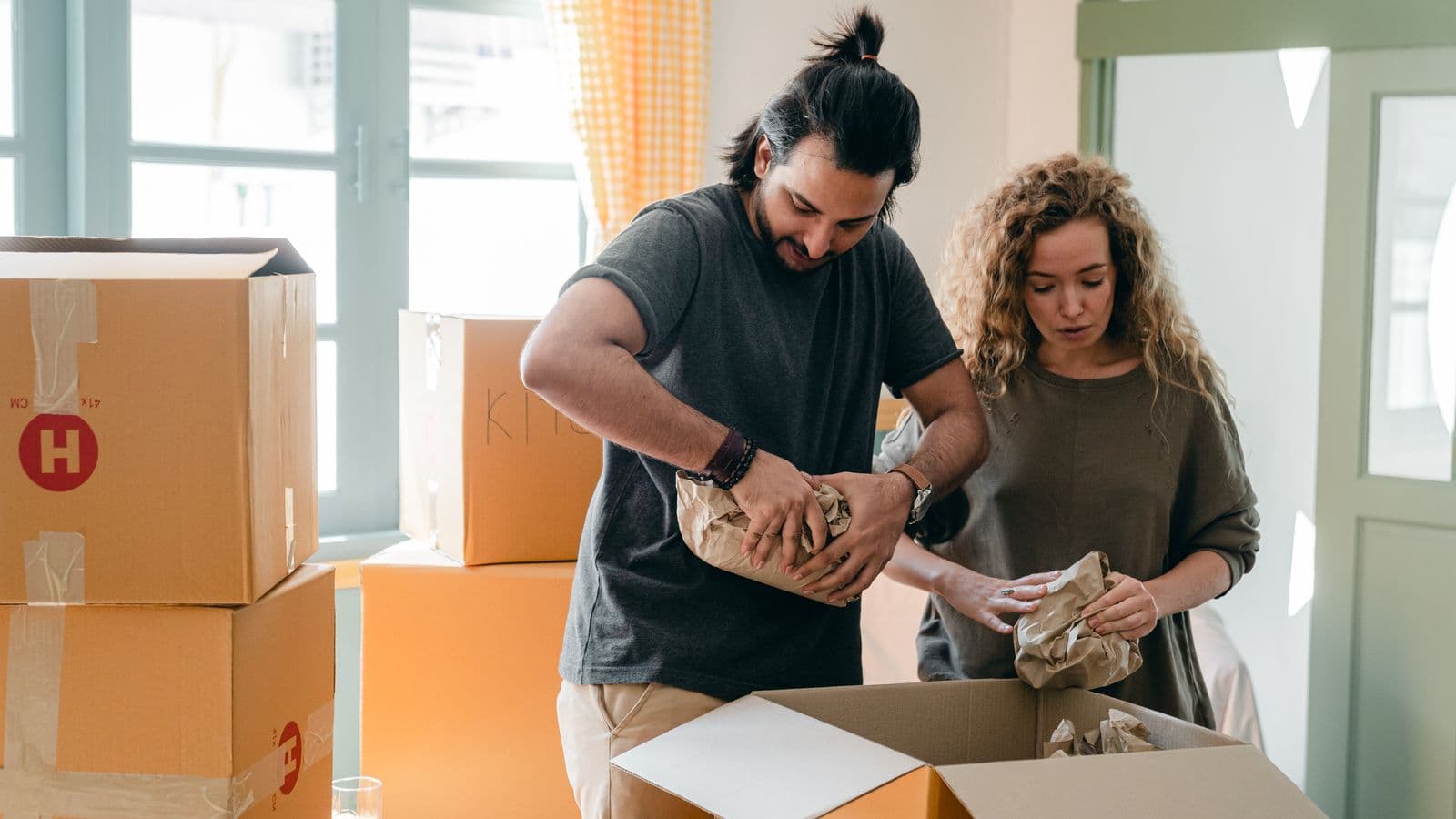 A couple wrapping household belongings beside moving boxes in a bright home